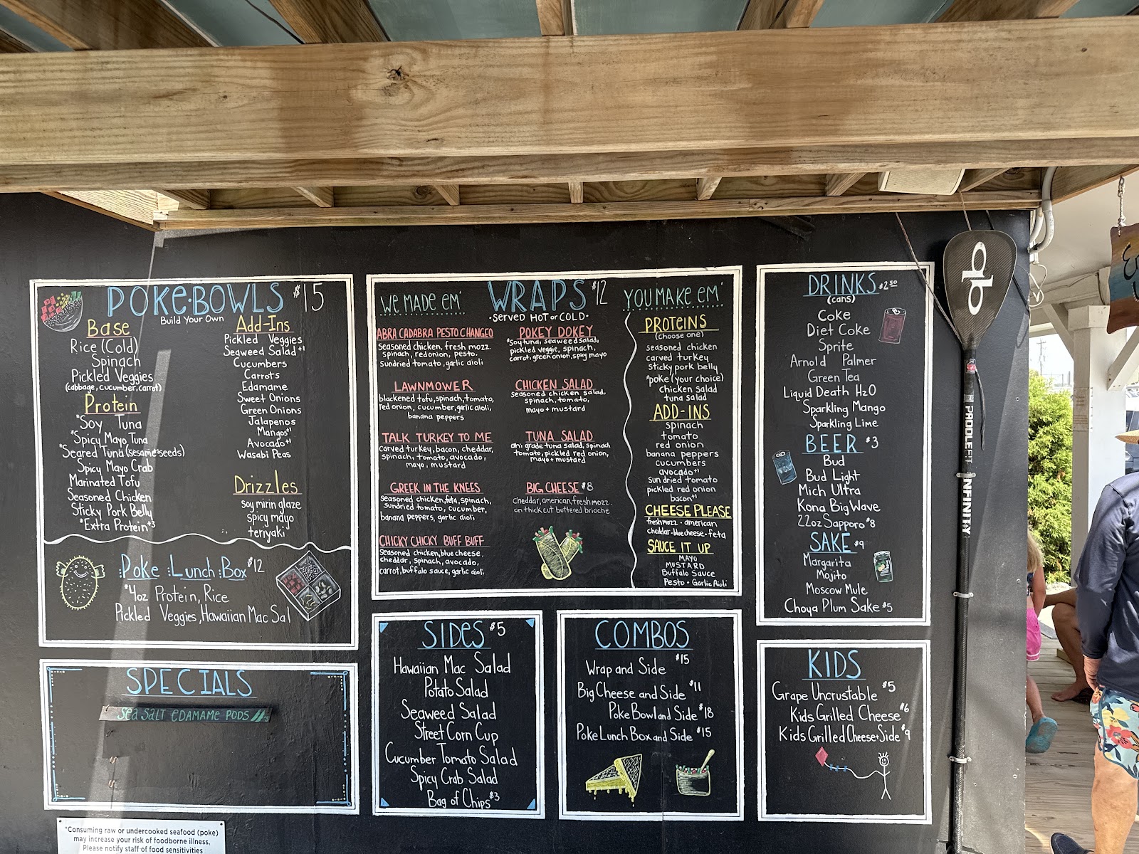 The Fast Fish hand-lettered chalkboard menu under the wooden awning at the Surf City stand, listing poke bowls, wraps, sides, combos, drinks, beer and sake.